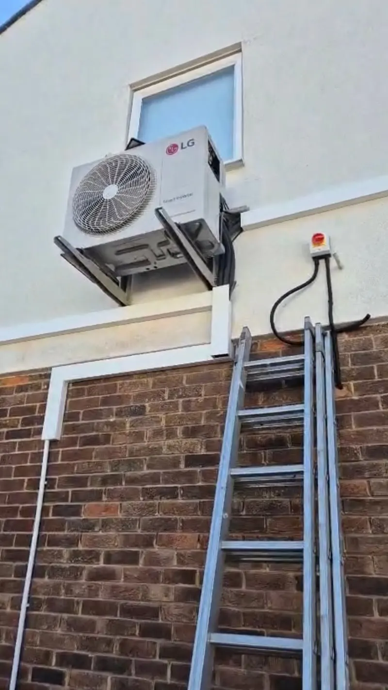 Outdoor air conditioning unit on wall brackets above brick, with isolator switch and pipework in white trunking