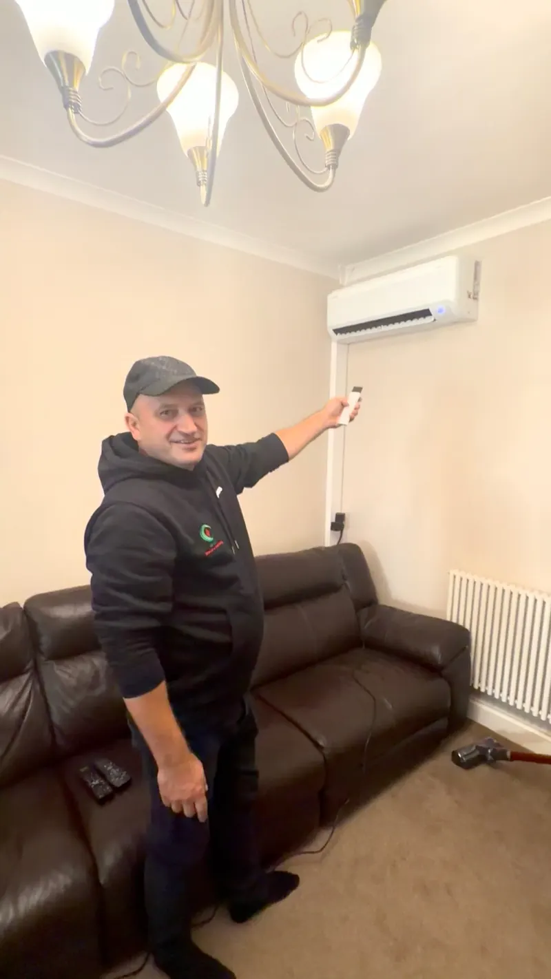 Engineer holding the controller beside a wall-mounted indoor unit above a sofa, next to an existing radiator