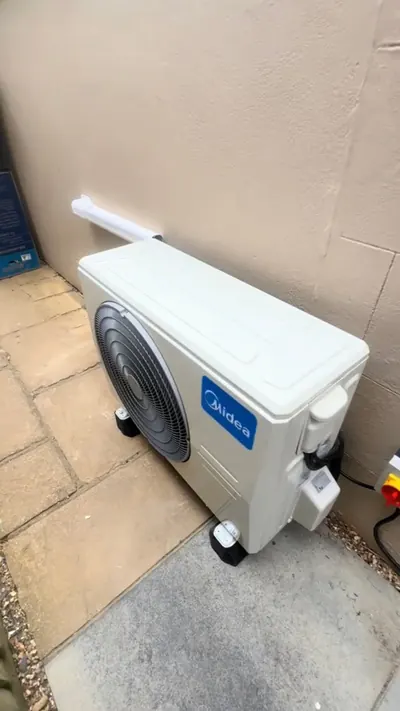 Midea outdoor unit installed on paving against a rendered wall with condensate run